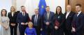Na zdjęciu jest grupa dziewięciu osób ( cztery uśmiechnięte kobiety w tym jedna kobieta  na wózku inwalidzkim oraz pięciu mężczyzn w garniturach).Grupa dziewięciu osób znajduje się w pokoju,a za ich plecami jest flaga Unii Europejskiej niebieska z żółtymi gwiazdami oraz flaga Polski biało- czerwona. 